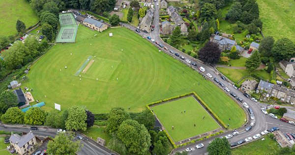 An aerial photograph of a green field, with a bowls lawn and trees at the bottom, bordered by a road with parked cars and trees lining a small stream.