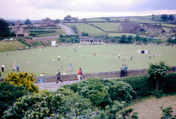 Many people in a field with a cricket pavilion in the background.