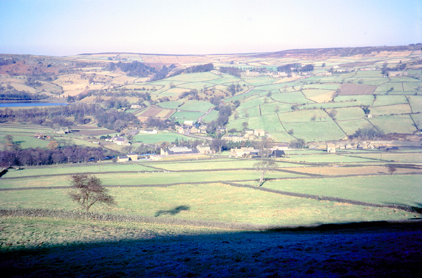 A patchwork of fields split by low stone walls across rolling hills, a small village at the bottom of a valley, and a body of water surrounded by trees to the left.