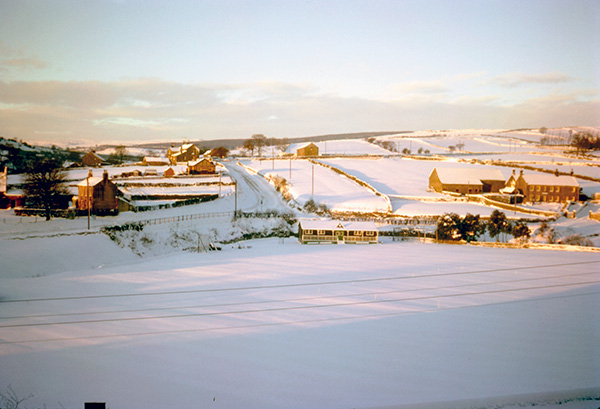 A white field, cricket pavilion, houses and outbuildings.